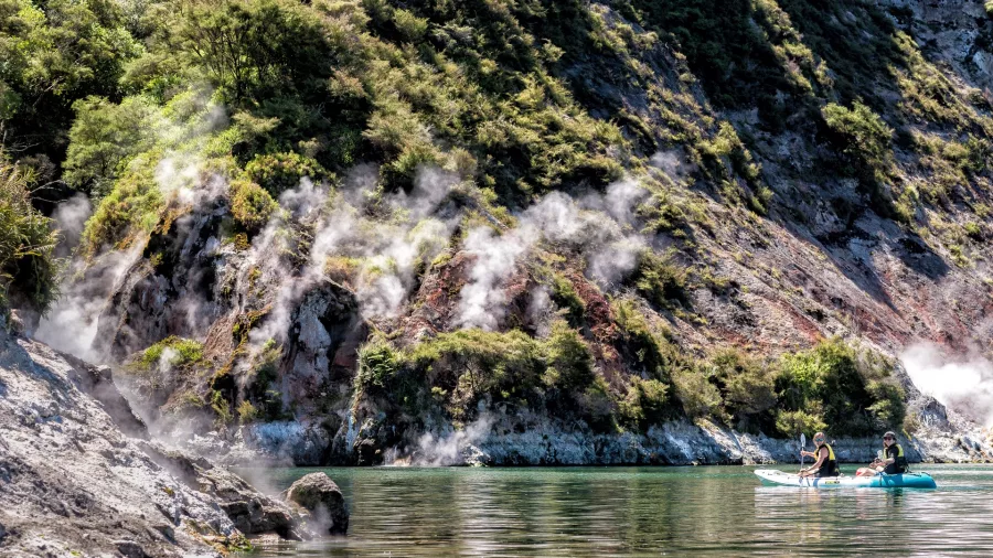 Kayakers passing steaming geothermal cliffs surrounded by lush native bush at Lake Rotomahana