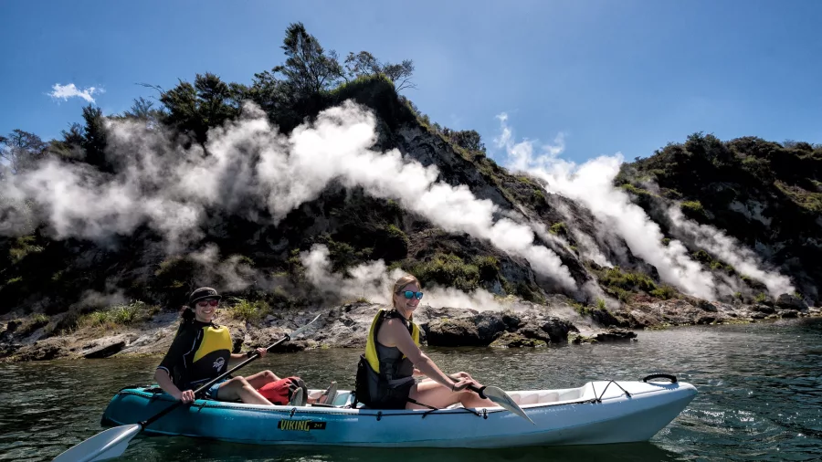 Two people kayaking beside geothermal steam vents rising from Lake Rotomahana's cliffs.