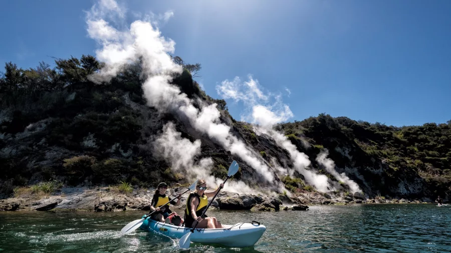 Two kayakers paddling past steaming cliffs on Lake Rotomahana in Waimangu Valley.
