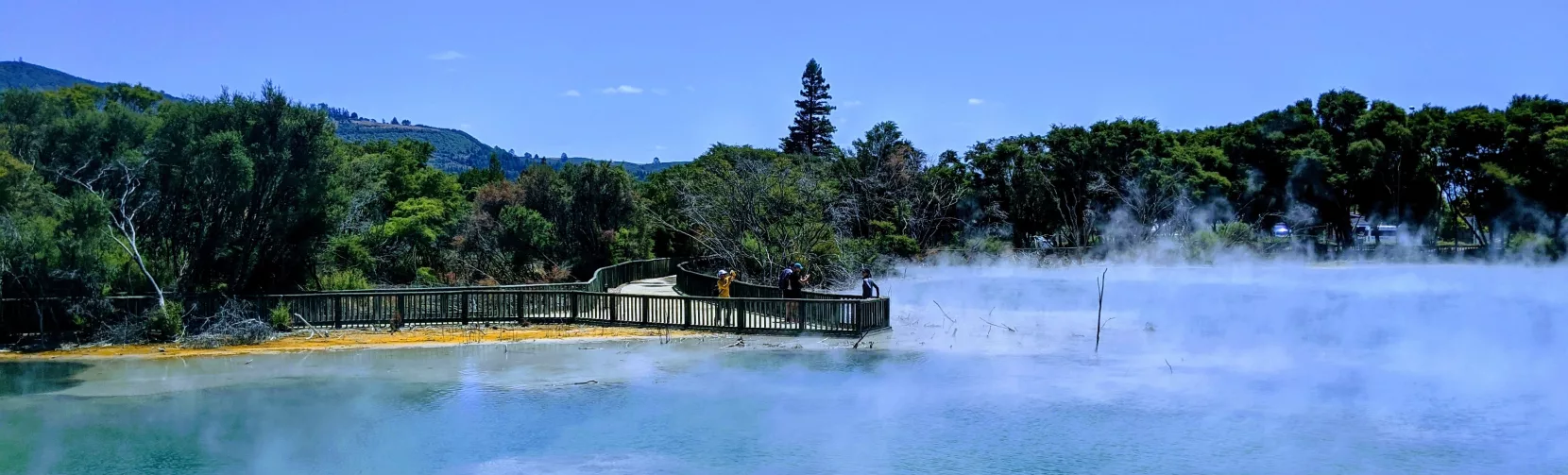 Steam rising from a pale blue geothermal lake with a boardwalk in Rotorua.