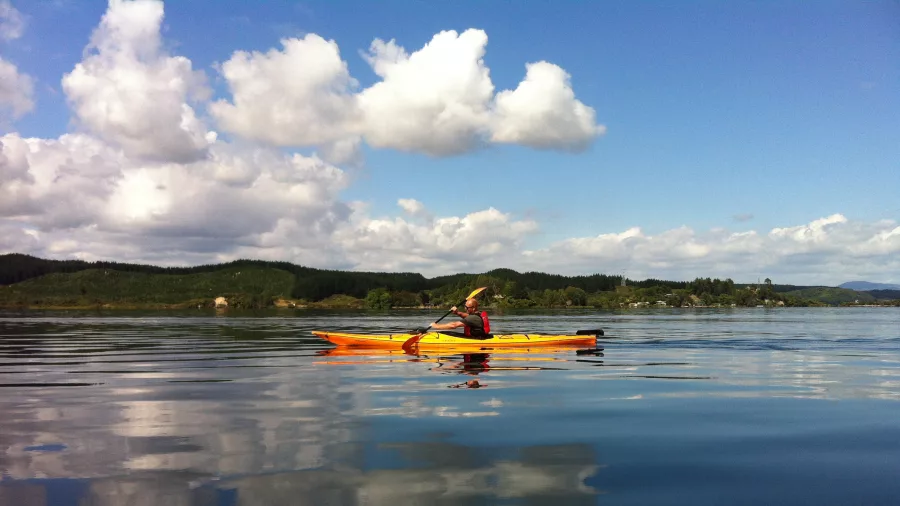 Solo paddler in a yellow kayak gliding across Lake Rotoiti with cloud reflections on the water.