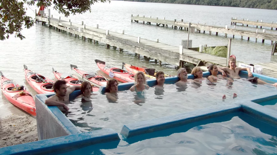 Group relaxing in a hot pool beside Lake Rotoiti with red kayaks and a wooden wharf in view.