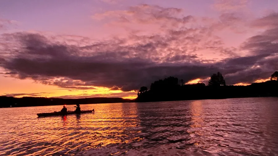 Silhouetted kayakers paddling Lake Rotoiti at sunset, glowing skies reflected in the water.
