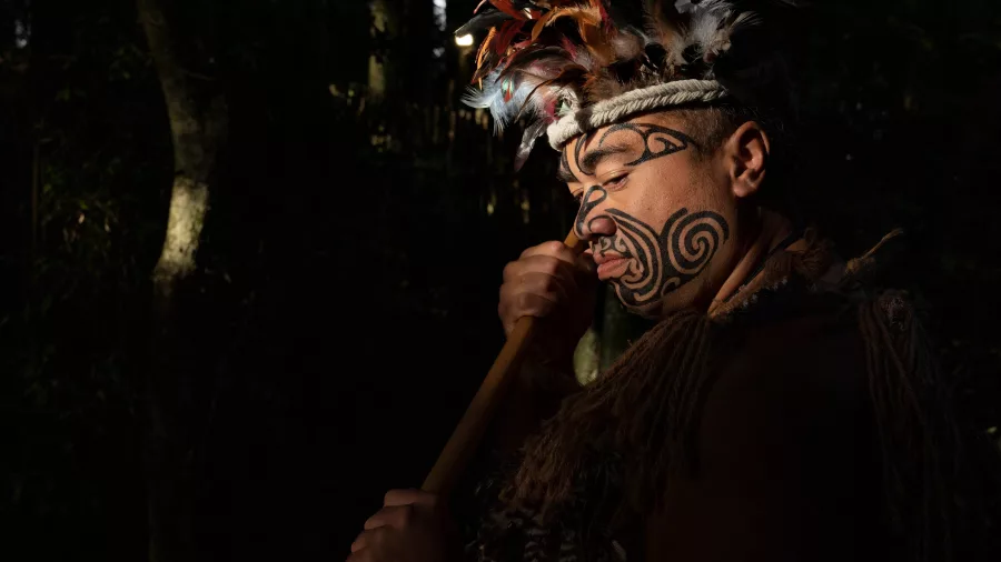 Māori man with tā moko (tattoo) and traditional attire holding a taiaha (club) at Te Pā Tū