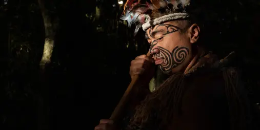 Māori man with tā moko (tattoo) and traditional attire holding a taiaha (club) at Te Pā Tū