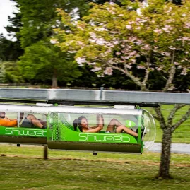 Shweeb Racer participants in motion pedalling past a lush grassy backdrop