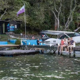 Group sitting on a wooden jetty beside a Volcanic Air floatplane at Lake Rotoiti Hot Pools