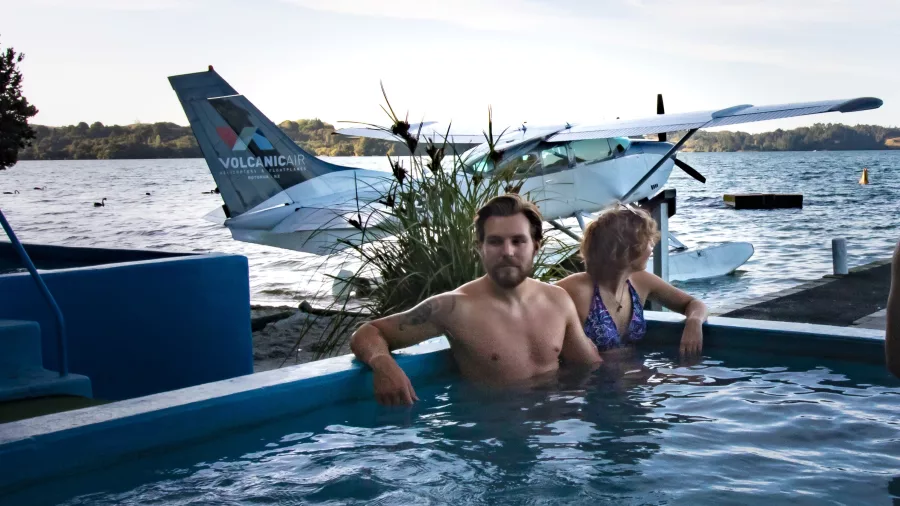 Couple relaxing in a hot pool with a Volcanic Air floatplane parked at Lake Rotoiti