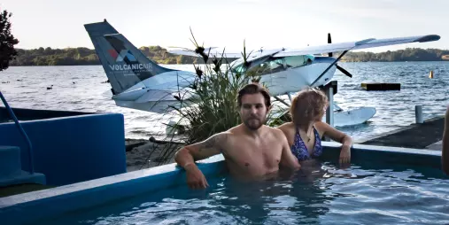 Couple relaxing in a hot pool with a Volcanic Air floatplane parked at Lake Rotoiti