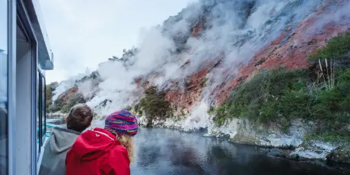 Passengers watching geothermal steam rise from red cliffs during a Waimangu boat cruise