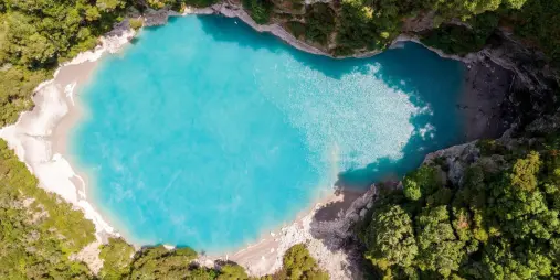 Aerial view of vivid blue Inferno Crater surrounded by native bush in Waimangu Volcanic Valley