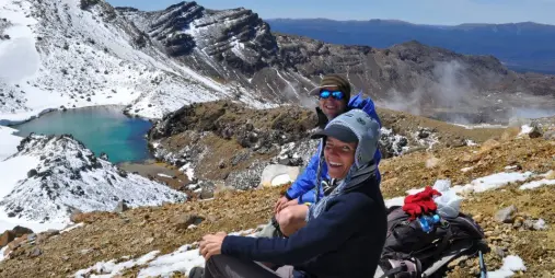 Two hikers resting on a snow-covered trail overlooking Emerald Lake in Tongariro National Park