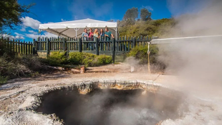 Visitors observing a steaming geothermal pool at Te Puia in Rotorua, New Zealand, with native landscape in the background.