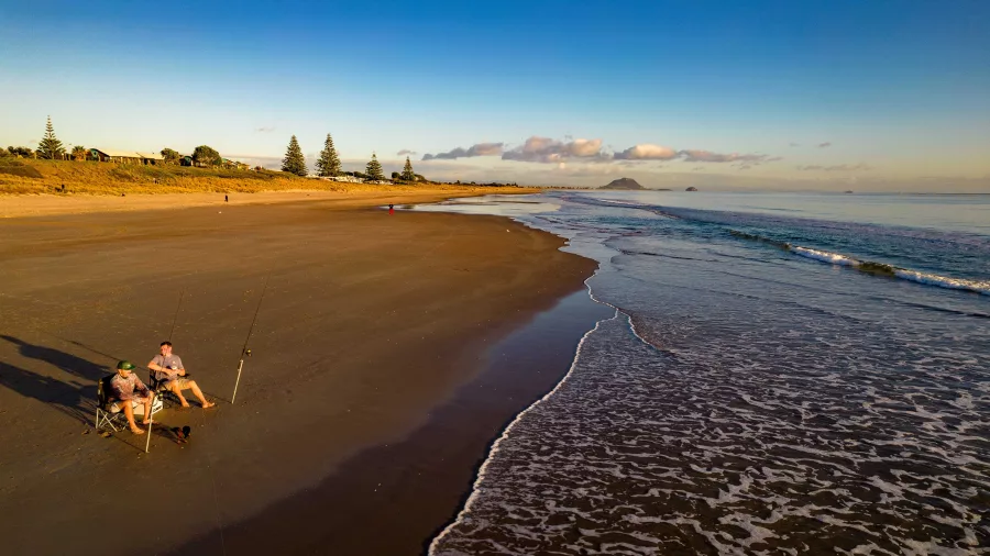 Two people fishing at sunrise on the sandy shoreline of Papamoa Beach, Bay of Plenty, New Zealand