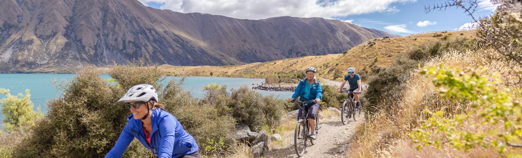Cyclists riding along a gravel track beside Lake Ohau with hills and clouds overhead.