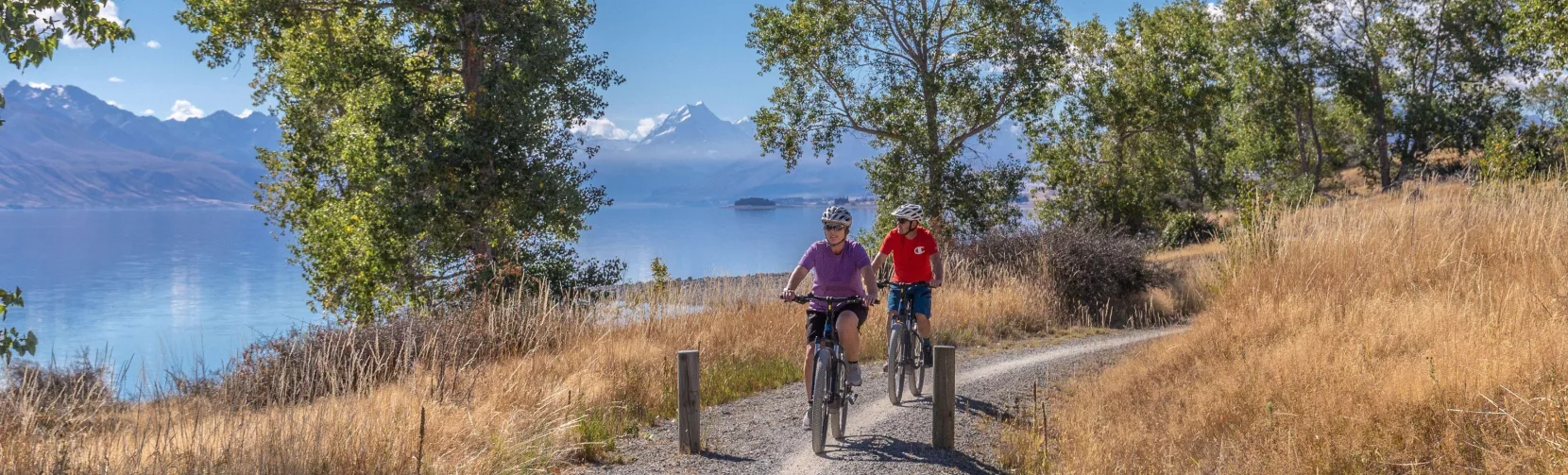 Two cyclists pedalling along a gravel path beside the brilliant blue waters of Lake Pukaki with mountains in the distance.