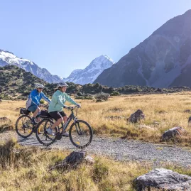 Two cyclists on a gravel trail with Aoraki/Mt Cook visible in the distance.