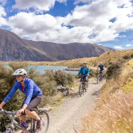 Cyclists riding along a gravel track beside Lake Ohau with hills and clouds overhead.