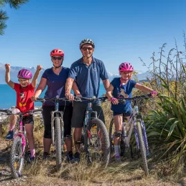 A family of four posing with bikes beside the bright turquoise waters of Lake Pukaki.