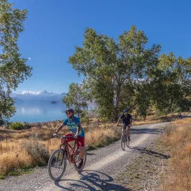 Cyclists on a gravel trail beside Lake Pukaki with mountains and trees in the background.