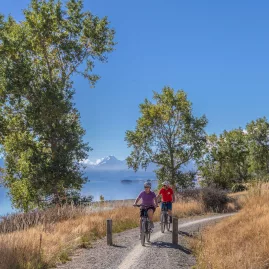 Two cyclists pedalling along a gravel path beside the brilliant blue waters of Lake Pukaki with mountains in the distance.