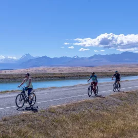 Cyclists riding alongside the turquoise Tekapo Canal on a sunny day with mountain views in the background.
