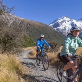 Couple riding bikes along a gravel trail with Aoraki/Mount Cook in the background.
