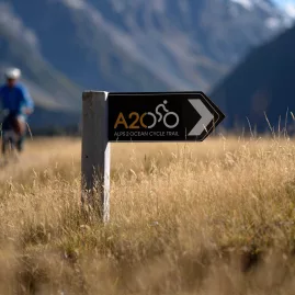 Alps 2 Ocean Cycle Trail directional sign with two cyclists in the background near Aoraki/Mount Cook.