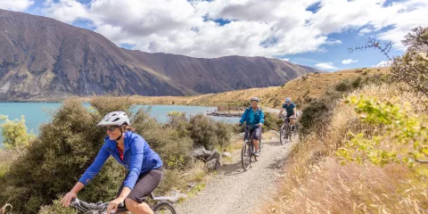 Cyclists riding along a gravel track beside Lake Ohau with hills and clouds overhead.