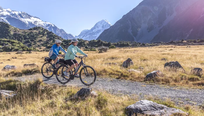 Two cyclists on a gravel trail with Aoraki/Mt Cook visible in the distance.