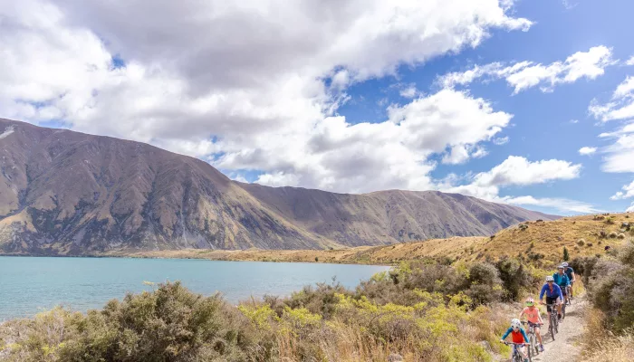 A group of cyclists, including children, riding next to the shoreline of Lake Ohau.