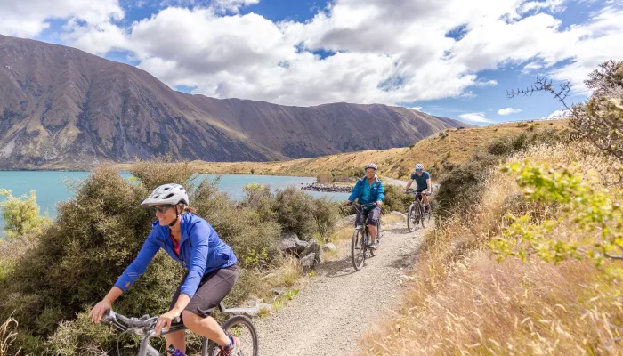 Cyclists riding along a gravel track beside Lake Ohau with hills and clouds overhead.