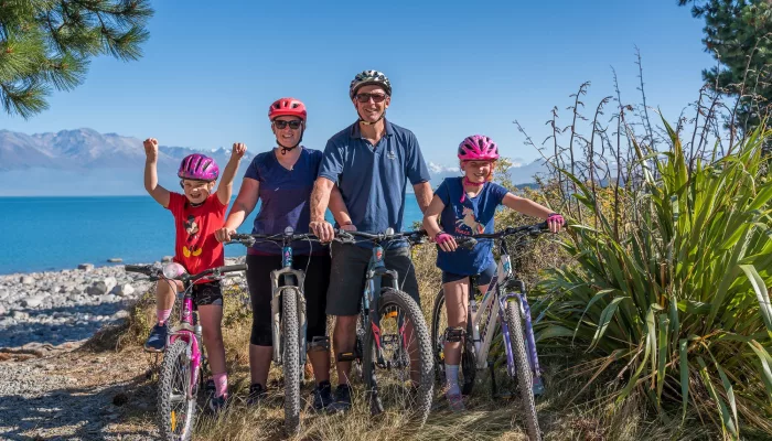 A family of four posing with bikes beside the bright turquoise waters of Lake Pukaki.