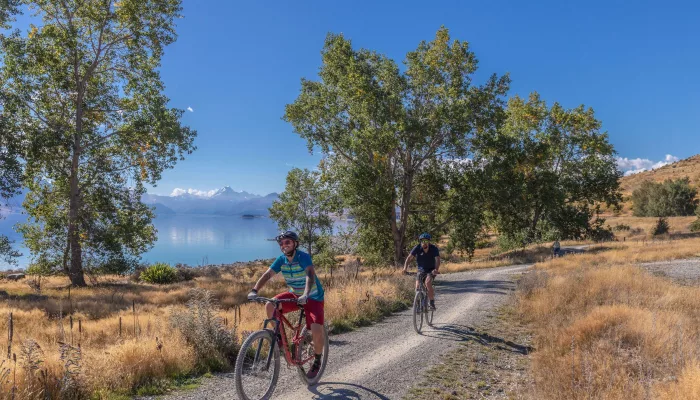Cyclists on a gravel trail beside Lake Pukaki with mountains and trees in the background.