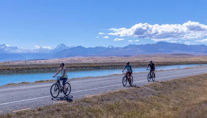 Cyclists riding alongside the turquoise Tekapo Canal on a sunny day with mountain views in the background.