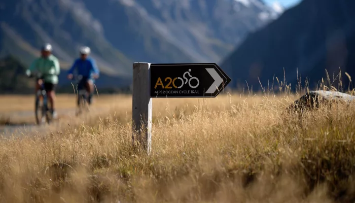 Alps 2 Ocean Cycle Trail directional sign with two cyclists in the background near Aoraki/Mount Cook.