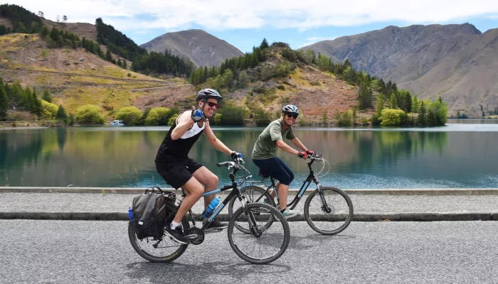 Two cyclists riding past a calm lake near Kurow on the Alps 2 Ocean Cycle Trail.