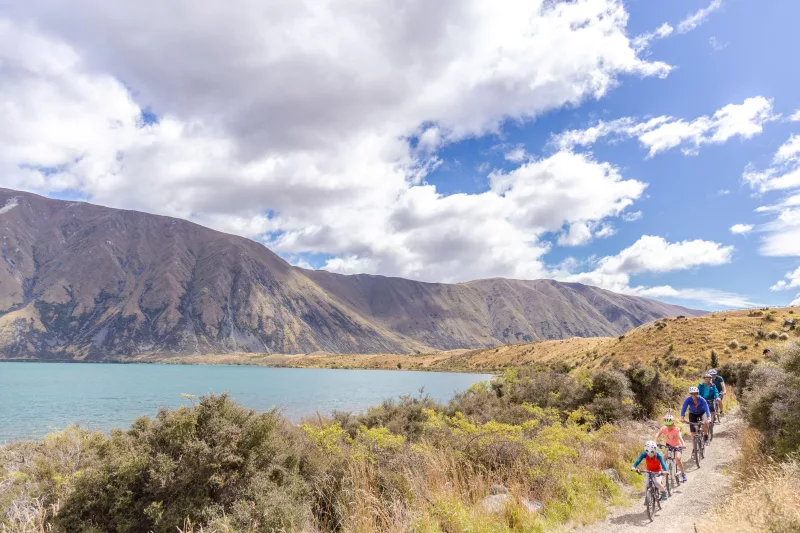 A group of cyclists, including children, riding next to the shoreline of Lake Ohau.