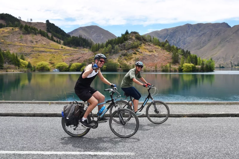 Two cyclists riding past a calm lake near Kurow on the Alps 2 Ocean Cycle Trail.