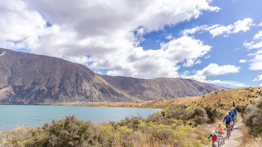 A group of cyclists, including children, riding next to the shoreline of Lake Ohau.
