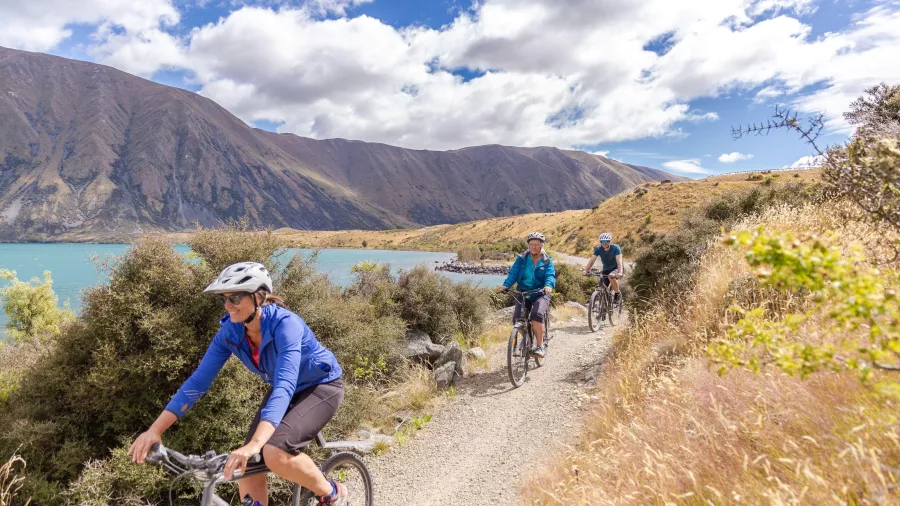 Cyclists riding along a gravel track beside Lake Ohau with hills and clouds overhead.