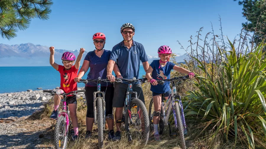 A family of four posing with bikes beside the bright turquoise waters of Lake Pukaki.