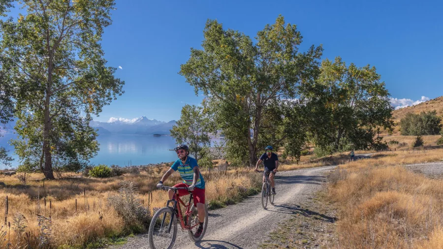 Cyclists on a gravel trail beside Lake Pukaki with mountains and trees in the background.