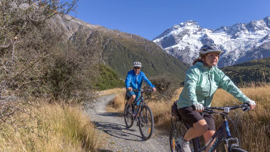 Couple riding bikes along a gravel trail with Aoraki/Mount Cook in the background.