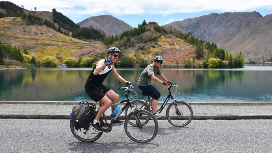 Two cyclists riding past a calm lake near Kurow on the Alps 2 Ocean Cycle Trail.