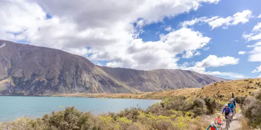A group of cyclists, including children, riding next to the shoreline of Lake Ohau.