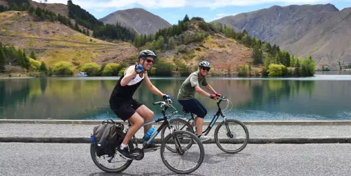 Two cyclists riding past a calm lake near Kurow on the Alps 2 Ocean Cycle Trail.