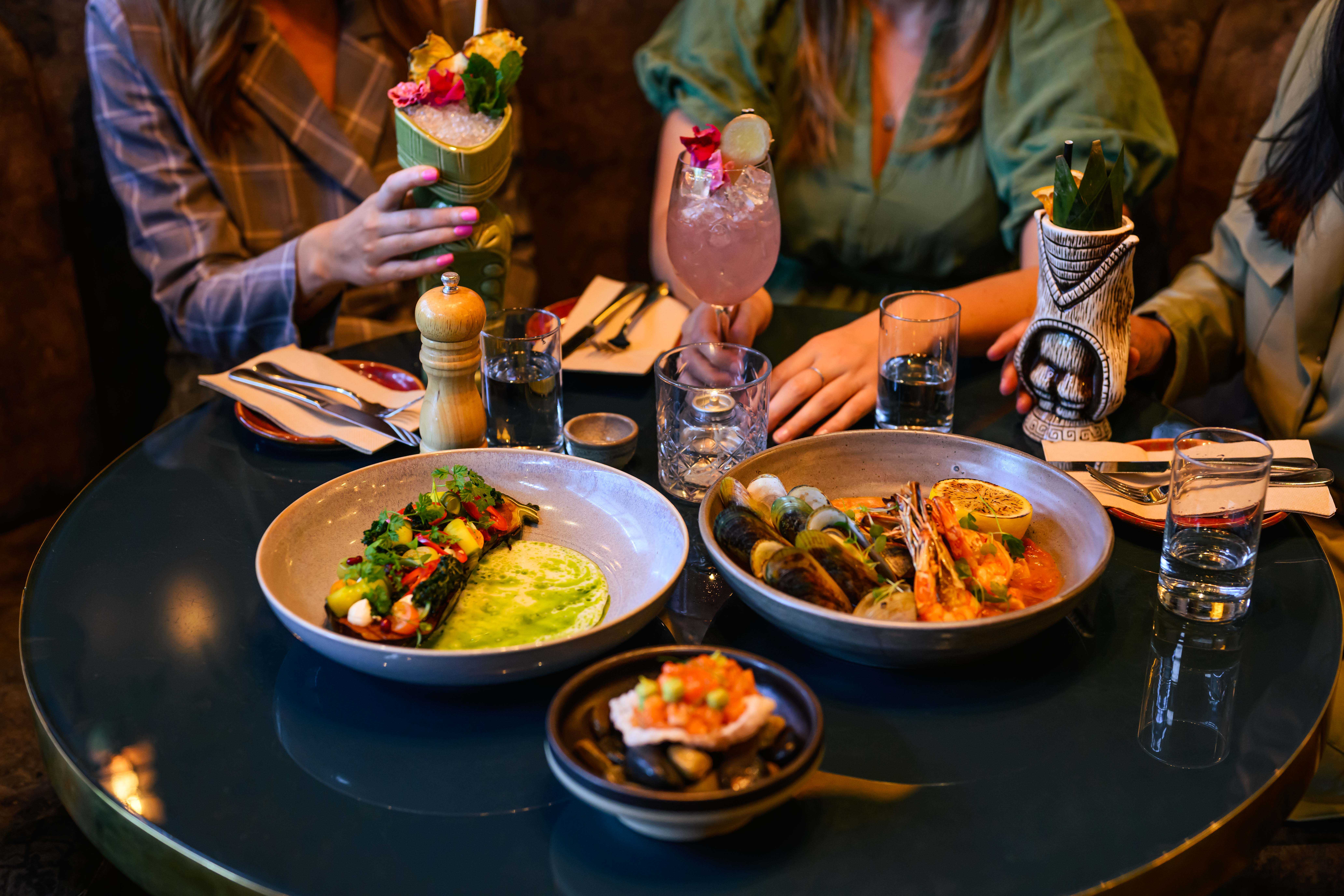 Three people dining at a warmly lit restaurant in Christchurch, New Zealand, with colourful cocktails, seafood dishes, shared plates, and water glasses on the table. Photo by Alistair Guthrie.