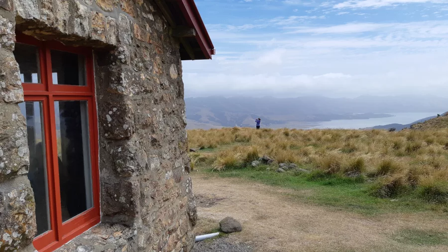 Stone wall and red window of Packhorse Hut with Lyttelton Harbour in the background