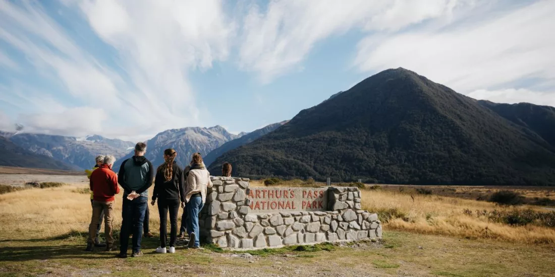 Group of visitors standing at the Arthur’s Pass National Park sign with mountain backdrop